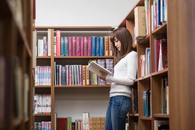 図書館の本棚にもたれて本を読む女子大生1